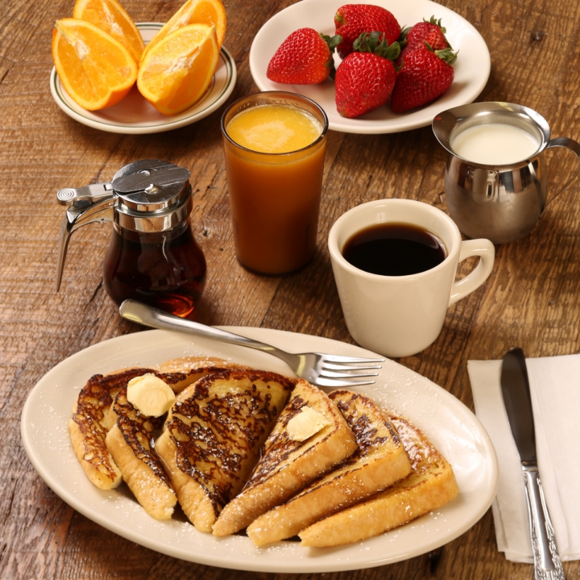 Image contexte Yami, table de petite déjeuner, avec des assiettes de fruits, un café, un verre de jus d’orange, un pot de lait et du pain perdu, sur une table en bois