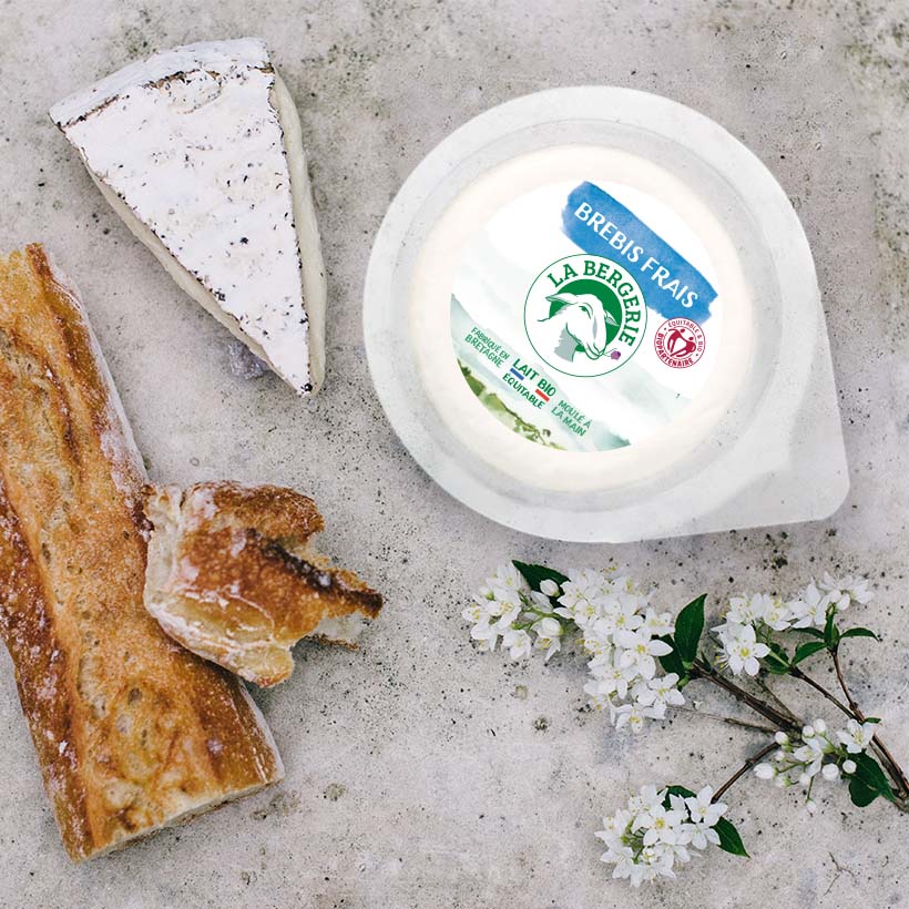 Instant de consommation brebis frais La Bergerie, sur un plan de travail, avec une part de fromage, un morceau de pain et des fleurs blanches